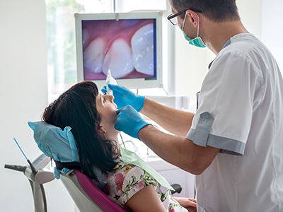 A dental professional is attending to a patient in a dental chair, with medical equipment and a monitor displaying a dental procedure being performed.