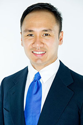 The image shows a man smiling at the camera, wearing a suit with a blue tie and a white shirt. He has short hair and is standing against a plain background.