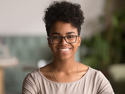 The image shows a woman with short hair, wearing glasses and a necklace, smiling and looking directly at the camera. She is dressed in a casual top and has a relaxed posture.