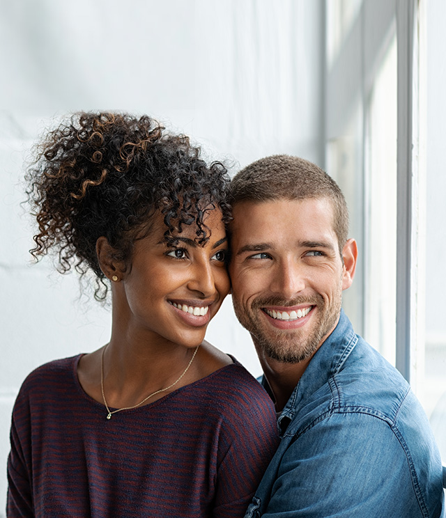 A young couple posing for a portrait with smiles, the man has short hair and is wearing a blue shirt, while the woman has curly hair and is in a dark top.