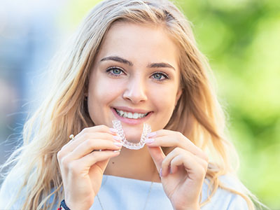 The image shows a smiling woman holding a clear braces appliance with her teeth.