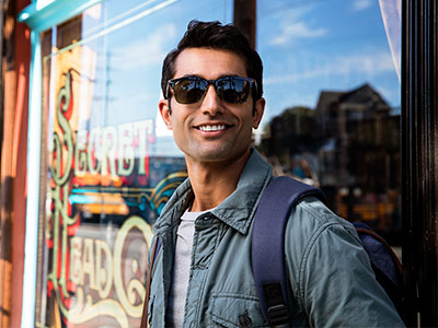 The image shows a man standing in front of a store window, wearing sunglasses and a backpack. He is smiling and appears to be posing for the photograph.