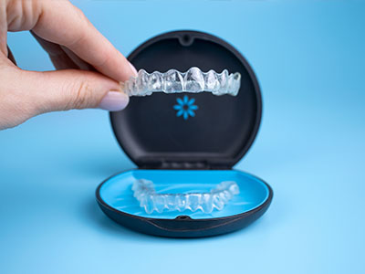 The image shows a hand holding an open case containing a set of clear, plastic dental aligners, with one aligner partially inserted into a blue mouthpiece.