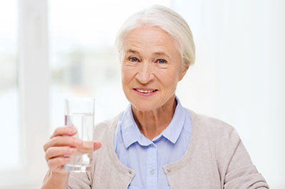 The image shows an elderly woman holding a glass of water, smiling at the camera.