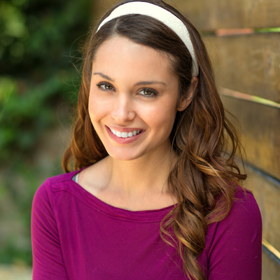 A woman with a bright smile, wearing a purple top and a headband, stands confidently against a wooden fence.