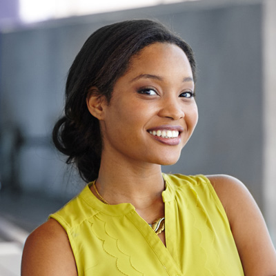 A young woman with a radiant smile, wearing a yellow top and standing against a backdrop of a building.