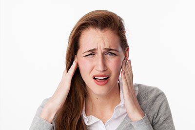 A woman with blonde hair, wearing a gray sweater and holding her hand to her ear, appears to be in pain or discomfort.