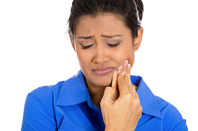 The image shows a woman with her eyes closed, holding her hand to her face in a gesture that could suggest she is experiencing pain or discomfort. She appears to be indoors and is wearing a light blue top.