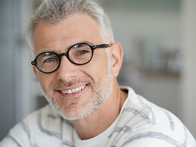 The image is a photograph of an older man with gray hair, wearing glasses and a white shirt. He has a beard and mustache, and appears to be smiling at the camera.