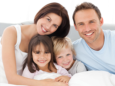 The image depicts a family of four, including two adults and two children, lying in bed with smiles on their faces.