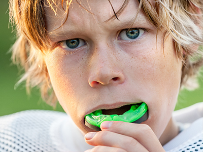 A young boy with blonde hair and blue eyes, wearing a football uniform, holds a green toothbrush in his mouth.