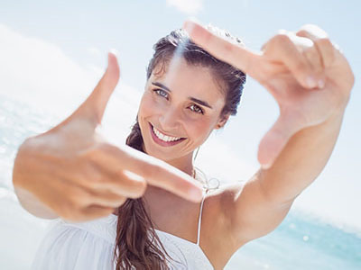 A young woman with long hair, smiling, holds up a hand to frame her face against the bright sky.